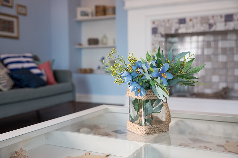 A cozy living room with a light blue color scheme featuring a glass vase wrapped in rope, filled with blue flowers and green foliage, placed on a glass coffee table. The background includes a grey couch with striped and solid pillows, built-in shelves with decorative items, and a fireplace with a tiled surround.