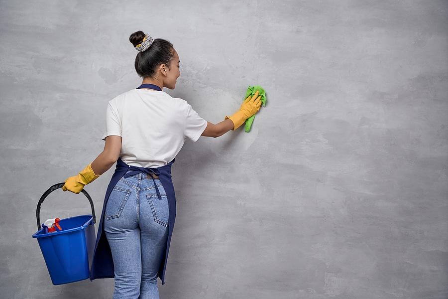 Young woman cleaning a gray wall with a green cloth while holding a blue bucket with cleaning supplies. She wears yellow rubber gloves, a white shirt, and an apron.